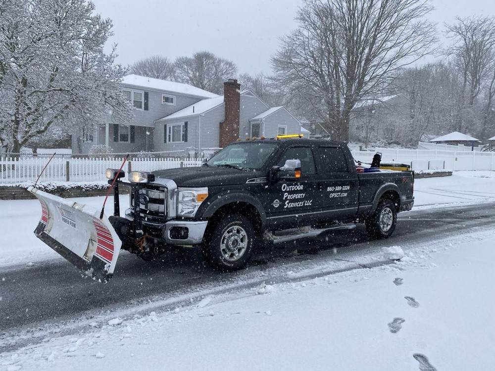 A lettered Outdoor Property Services plow truck on a snowy residential street with the snow still falling