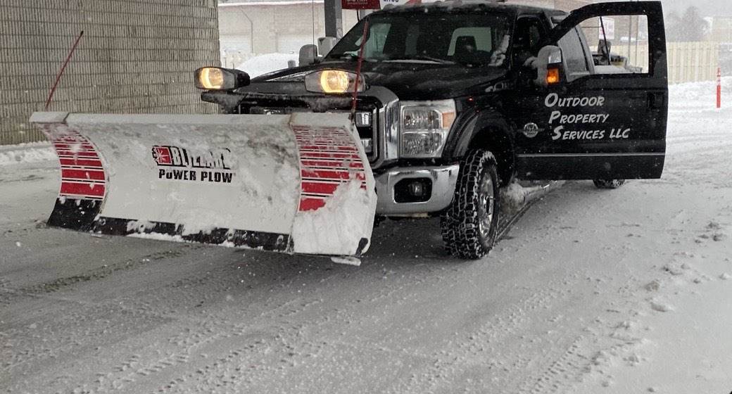 A lettered Outdoor Property Services plow truck clearing under the canopy of a fuel stop during a storm