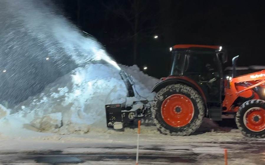 An Outdoor Property Services tractor with a snow blower throwing snow onto a pile in a lit commercial parking lot at night