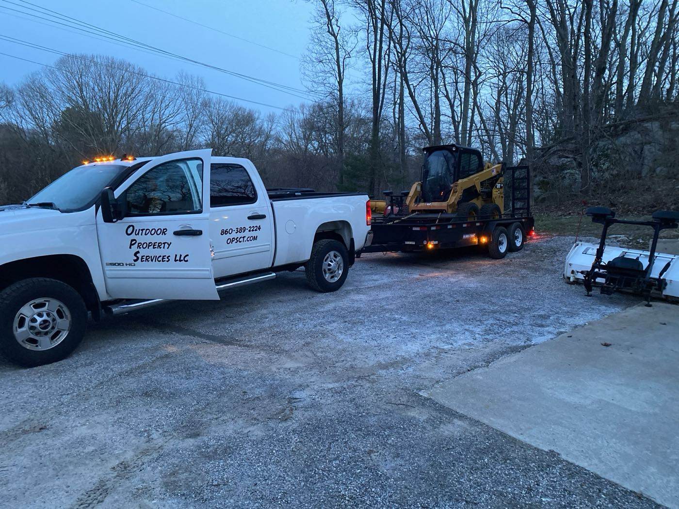 A lettered Outdoor Property Services truck towing the trailer with the tracked skid steer on it at dusk