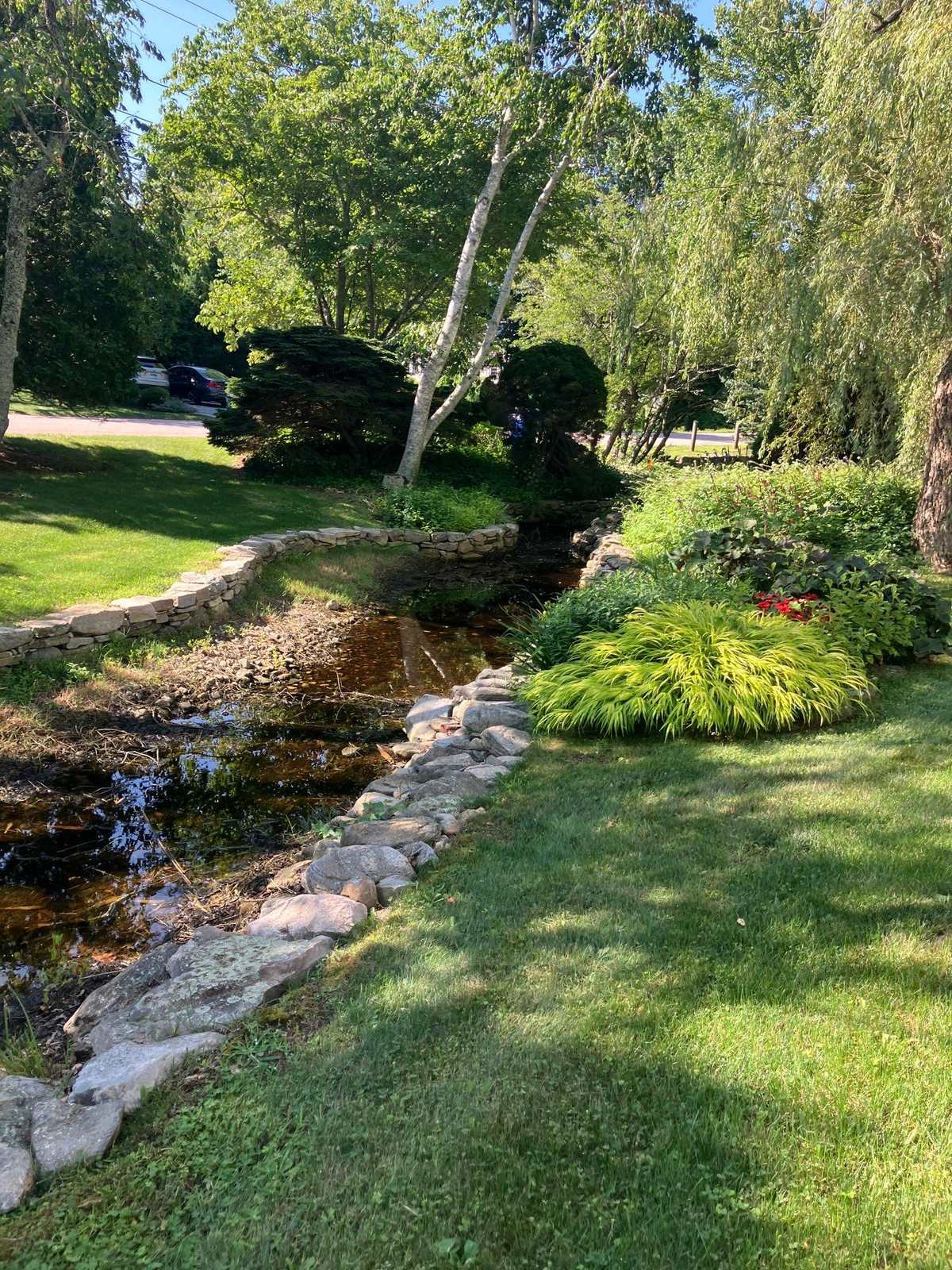A mown lawn and planted beds along a stone-lined stream on a Mystic, Connecticut property kept by Outdoor Property Services