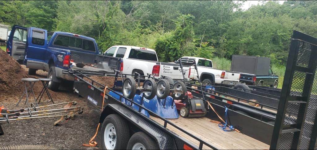 An Outdoor Property Services trailer loaded with commercial mowers, the pickups lined up behind it at the yard