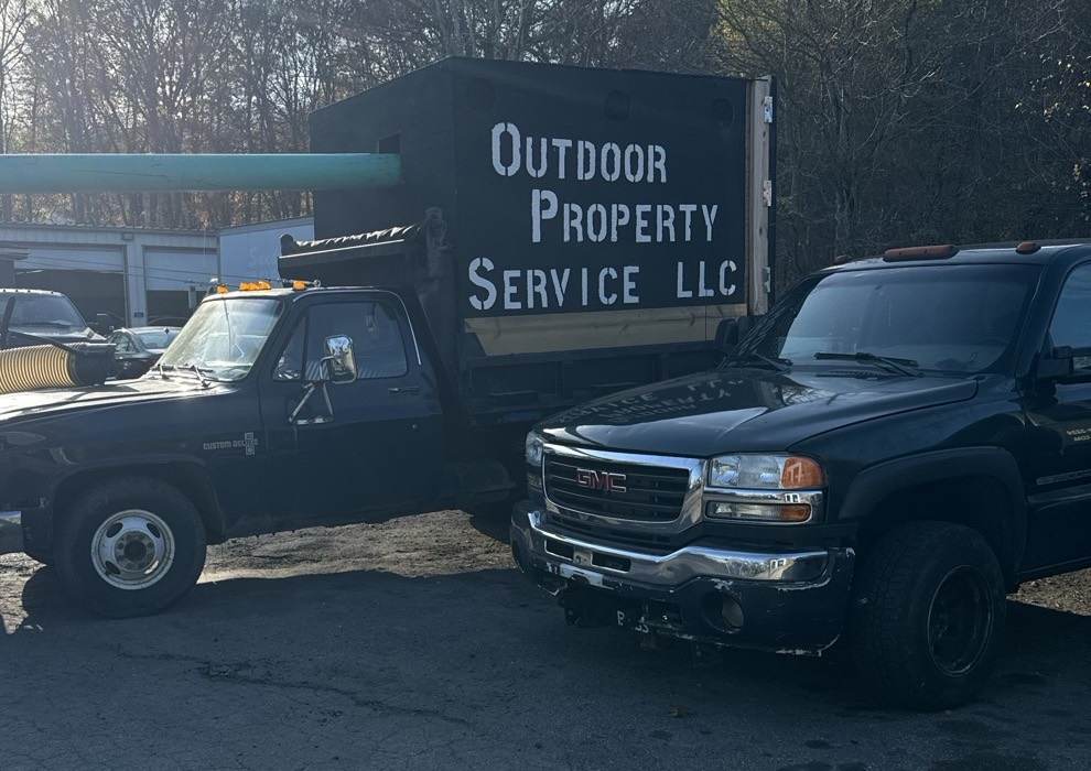 The lettered Outdoor Property Services leaf box on the chassis truck at the shop, with the loader hose beside it and a pickup alongside