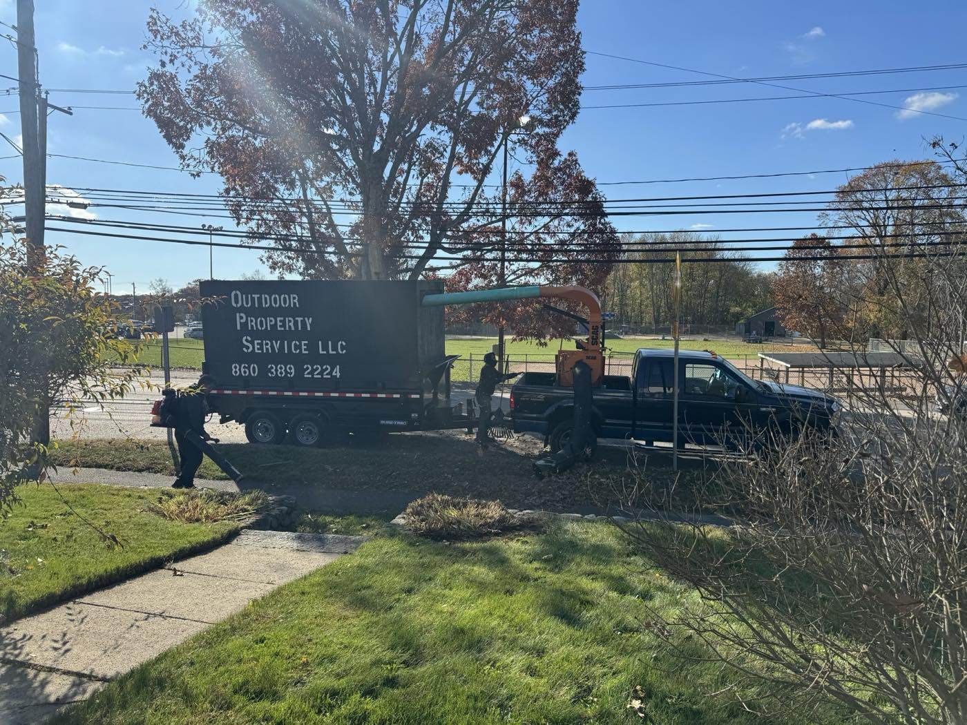 The lettered Outdoor Property Services leaf box trailer and truck loader at the curb, the crew raking leaves to the hose on a fall morning