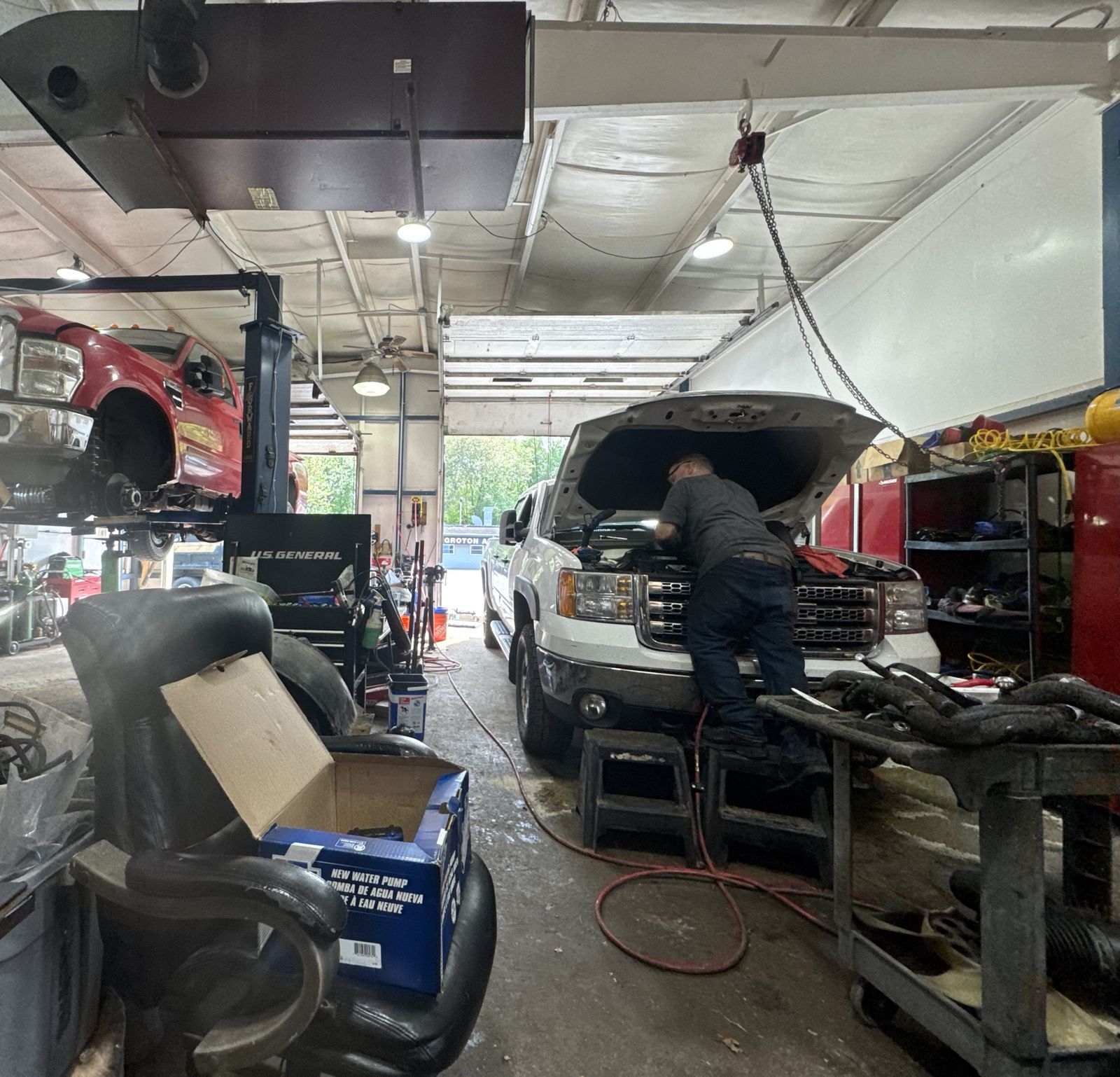 The Groton shop bay with both trucks in: the red F-350 up on the lift and the white GMC with its hood up and the mechanic at work