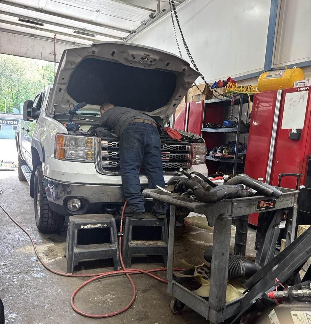 An Outdoor Property Services mechanic working under the hood of the white lettered GMC in the shop bay in Groton
