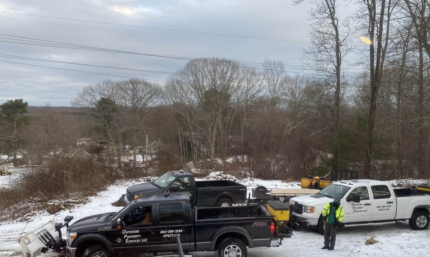 A lettered Outdoor Property Services plow truck with a tailgate spreader in the Groton yard before a storm