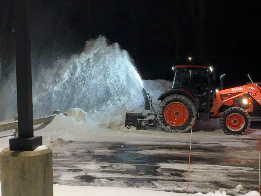 An Outdoor Property Services tractor with a snow blower throwing snow onto a pile in a lit commercial lot at night